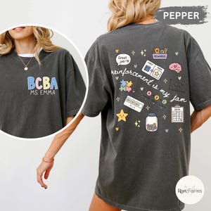 May include: A dark gray t-shirt with the text "BCBA MS. EMMA" in pastel colors on the front. The back features colorful graphics and the phrase "reinforcement is my jam."