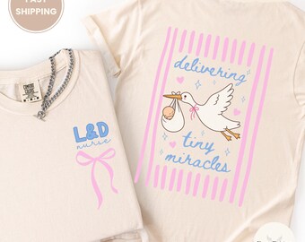 May include: Two ivory-colored t-shirts. One features "L&D nurse" in blue with a pink bow. The other has a stork carrying a baby with the words "delivering tiny miracles" in pink and blue.