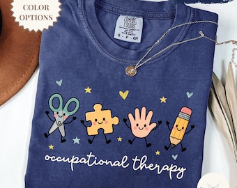 May include: A navy blue t-shirt featuring a graphic print of a smiling pair of scissors, a puzzle piece, a hand, and a pencil. The text "occupational therapy" is printed in white below the characters, surrounded by small yellow and blue stars and hearts.