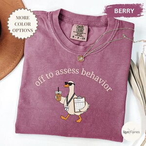 May include: A berry-colored crewneck t-shirt featuring a graphic of a goose wearing sunglasses, holding a coffee cup and a clipboard, with the text "off to assess behavior" printed above. The shirt has a Comfort Colors brand label at the neck.