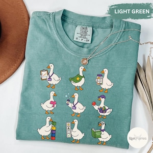 May include: A light green short-sleeve t-shirt featuring a graphic print of nine white geese in various roles. The geese are depicted with items including a clipboard, a star cape, a jar labeled Tiny Wins, a heart, bubbles, a tool belt, blocks, and a book.