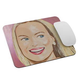 Könnte beinhalten: Ein rechteckiges Mauspad mit einem gemalten Porträt einer lächelnden Frau mit blonden Haaren und blauen Augen. Das Mauspad hat einen rosa Hintergrund und eine weiße Computermaus daneben.