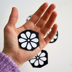 May include: A hand holding a black and white paper flower with a white center. There are two more paper flowers in the background.