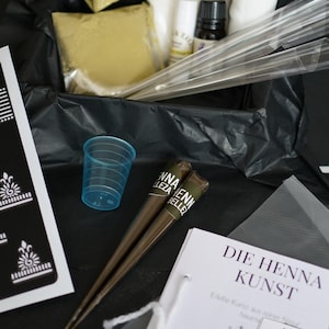 Puede incluir: Una caja negra llena de suministros de henna, incluyendo conos de henna, plantillas, una taza pequeña y un folleto titulado "Die Henna Kunst".