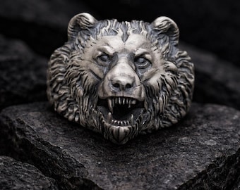 May include: A silver-toned metal ring featuring a detailed, three-dimensional bear head design. The bear is depicted with an open mouth, bared teeth, and textured fur detailing across the face and ears, finished with a dark, oxidized patina.