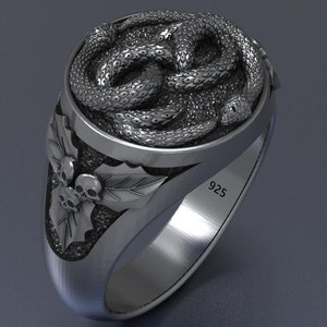 Può includere: Un anello d'argento con un designo dettagliato di serpenti intrecciati sulla parte superiore. La fascia dell'anello è decorata con una foglia e tre teschi. L'anello è contrassegnato dal numero "925" sul lato.