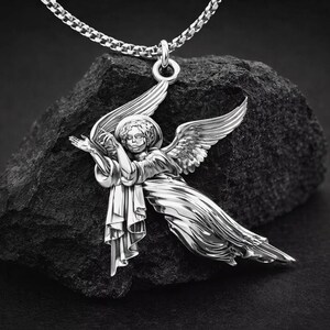 Puede incluir: Colgante de &aacute;ngel plateado con alas extendidas y un halo, suspendido de una cadena. El colgante detallado se encuentra sobre un fondo oscuro y texturizado. El &aacute;ngel est&aacute; representado con una t&uacute;nica fluida.