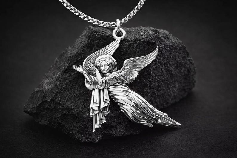 Puede incluir: Colgante de &aacute;ngel plateado con alas extendidas y un halo, suspendido de una cadena. El colgante detallado se encuentra sobre un fondo oscuro y texturizado. El &aacute;ngel est&aacute; representado con una t&uacute;nica fluida.