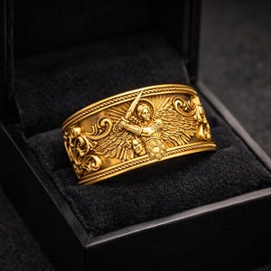 Puede incluir: Un anillo dorado con un dise&ntilde;o intrincado que presenta una figura alada sosteniendo una espada, colocado en una caja de joyer&iacute;a negra. El anillo tiene tallas detalladas y se muestra sobre una superficie de terciopelo negro.