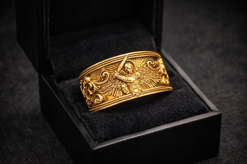 Puede incluir: Un anillo dorado con un dise&ntilde;o intrincado que presenta una figura alada sosteniendo una espada, colocado en una caja de joyer&iacute;a negra. El anillo tiene tallas detalladas y se muestra sobre una superficie de terciopelo negro.