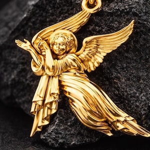 Puede incluir: Un collar con colgante de &aacute;ngel dorado. El colgante presenta un &aacute;ngel con alas extendidas y ropajes fluidos. El collar se muestra sobre una superficie oscura y texturizada, resaltando los intrincados detalles de la joya.