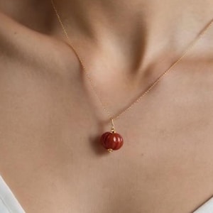 Peut inclure: Un collier en or avec un petit pendentif rond rouge. Le pendentif a une surface texturée et est suspendu à une fine chaîne en or. Le collier est porté sur un haut blanc.