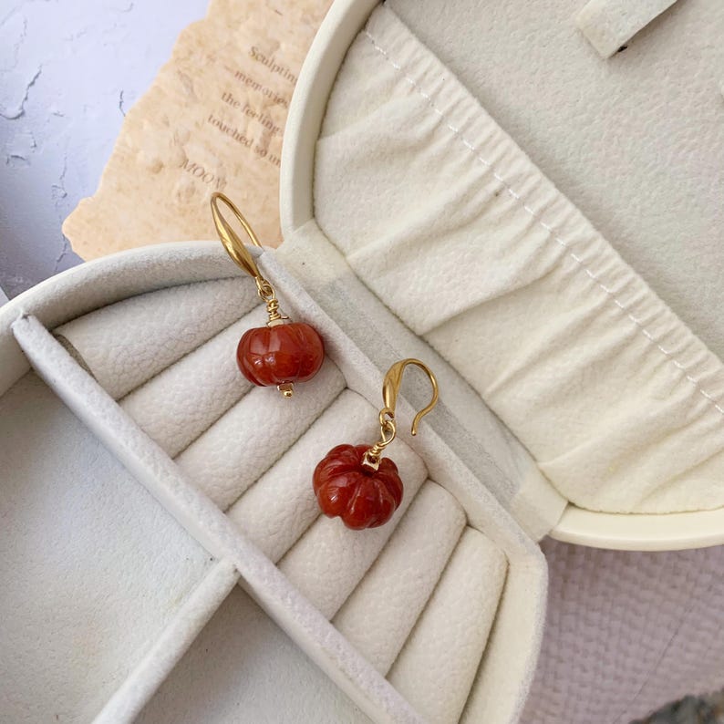 May include: A pair of gold-colored earrings featuring carved, reddish-orange pumpkin charms. The earrings are displayed inside an open, white jewelry box. The pumpkin charms have detailed ridges and are suspended from gold-colored hooks.