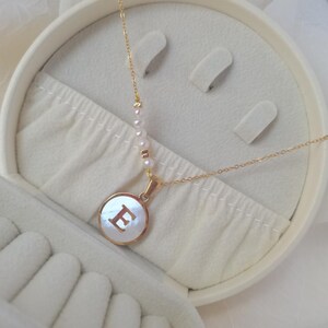 May include: A gold necklace with a pearl chain and a round pendant featuring the letter "E" in a white shell.