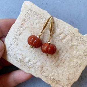 May include: A pair of pumpkin-shaped earrings. The earrings are a deep reddish-orange color, with gold-colored hooks and accents. The pumpkins have a carved texture. The earrings are displayed on a vintage paper with text.