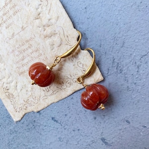 May include: A pair of gold-colored drop earrings featuring carved, reddish-orange pumpkin-shaped beads. The earrings are displayed on a piece of aged paper with handwritten text, set against a blue-gray background. The earrings have a delicate, autumnal aesthetic.