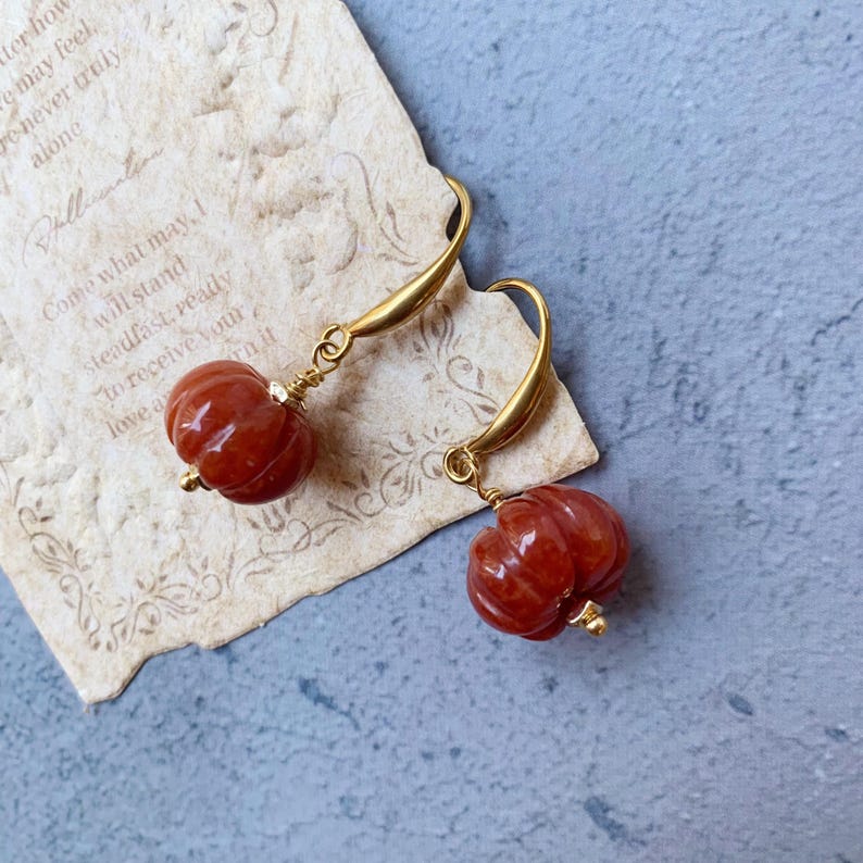 May include: A pair of gold-colored drop earrings featuring carved, reddish-orange pumpkin-shaped beads. The earrings are displayed on a piece of aged paper with handwritten text, set against a blue-gray background. The earrings have a delicate, autumnal aesthetic.