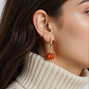 May include: A gold-colored earring with a small, orange pumpkin charm. The pumpkin has a ribbed texture and a small stem. The earring is hanging from a gold hook. The background is blurred, showing a person wearing a cream-colored sweater.