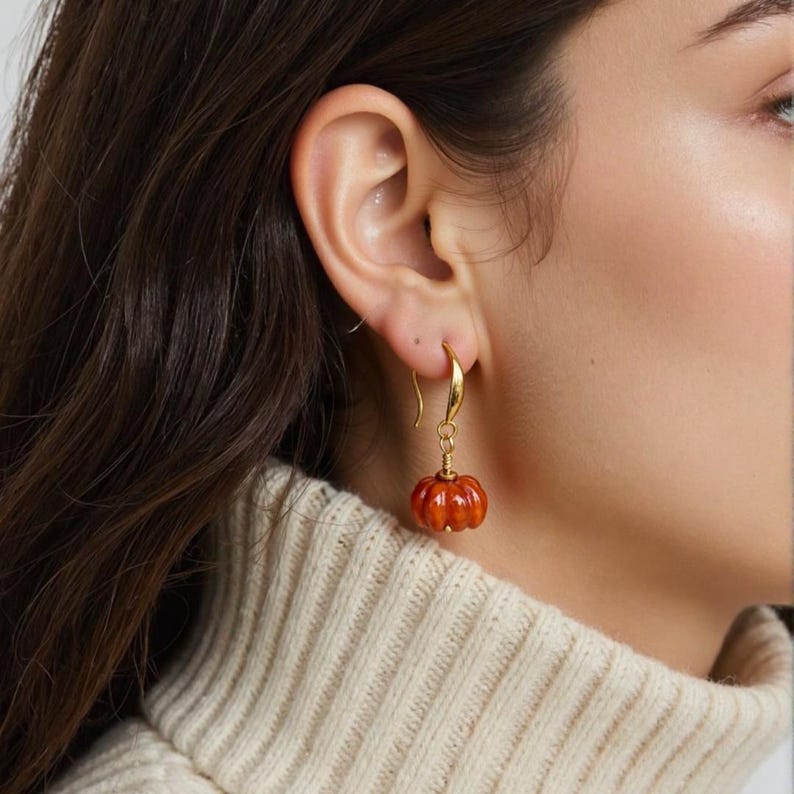May include: A gold-colored earring with a small, orange pumpkin charm. The pumpkin has a ribbed texture and a small stem. The earring is hanging from a gold hook. The background is blurred, showing a person wearing a cream-colored sweater.