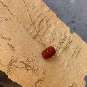 May include: A gold-colored necklace with a small, reddish-brown pumpkin pendant. The pendant is intricately detailed. The necklace rests on a piece of paper with text that reads "Dusk Dancing" and "Moonlight in a pocket."