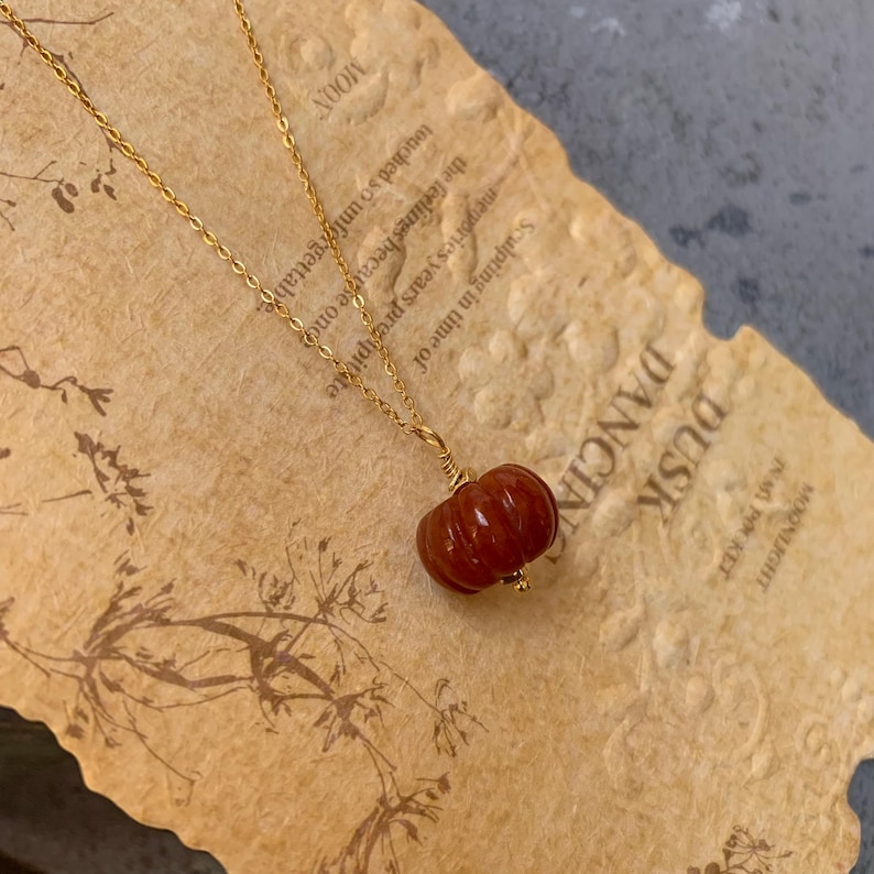 May include: A gold-colored necklace with a small, reddish-brown pumpkin pendant. The pendant is intricately detailed. The necklace rests on a piece of paper with text that reads "Dusk Dancing" and "Moonlight in a pocket."