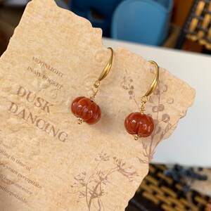 May include: A pair of gold-tone earrings featuring small, carved, reddish-brown pumpkin-shaped beads. The earrings are suspended from curved gold hooks. The text "MOONLIGHT IN MY POCKET" and "DUSK DANCING" is visible on the background.