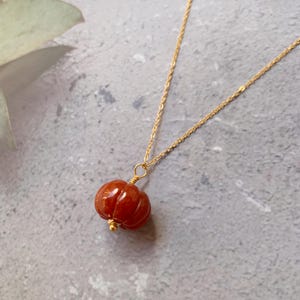 May include: A gold-colored chain necklace with a pendant shaped like a small, red-orange pumpkin. The pumpkin pendant has vertical grooves and a small gold bead at the bottom. The necklace is displayed on a gray surface.