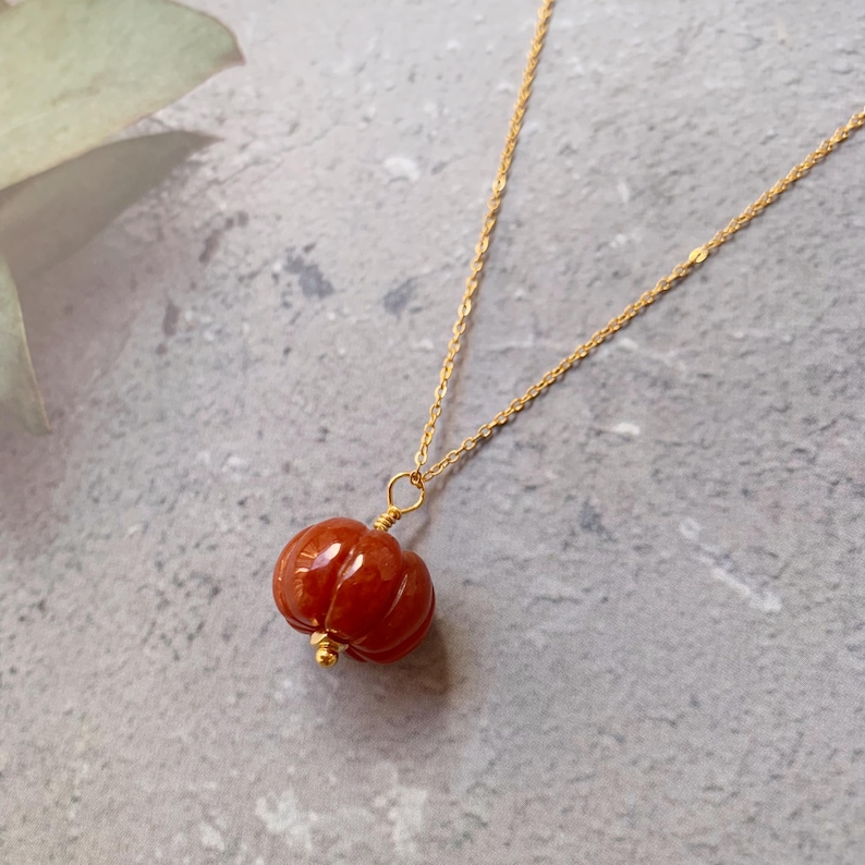 May include: A gold-colored chain necklace with a pendant shaped like a small, red-orange pumpkin. The pumpkin pendant has vertical grooves and a small gold bead at the bottom. The necklace is displayed on a gray surface.