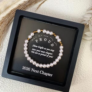 May include: A pearl bracelet with gold beads is presented on a black surface. The text reads "PROUD", "Shine bright as you step into your next chapter. We are so proud of you!" and "2026 Next Chapter". The bracelet is a delicate piece.