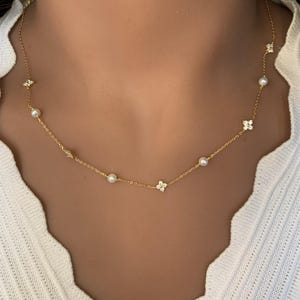 May include: A gold chain necklace with small pearl accents and delicate flower-shaped charms. The necklace is on a model's neck.