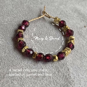May include: A gold-toned ring with deep red garnet beads and gold accents. The ring is inscribed with the words "Mary & David" and the phrase "A secret only you share, spelled in garnet and love."
