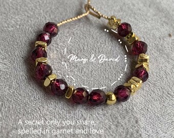 May include: A gold-toned ring with deep red garnet beads and gold accents. The ring is inscribed with the words "Mary & David" and the phrase "A secret only you share, spelled in garnet and love."