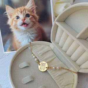 May include: A gold chain bracelet with a round charm featuring a black and white photo of a cat. The charm is surrounded by small white pearls. The bracelet is displayed in a white jewelry box.