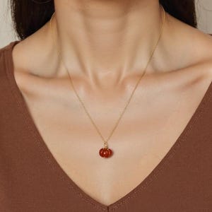 May include: A gold-colored necklace with a small, round, red pendant. The pendant has a textured surface and is suspended from a delicate chain. The necklace is worn around the neck.