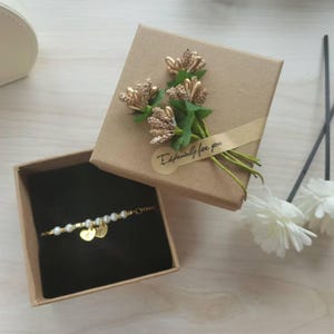 May include: A gold bracelet with pearls and two heart-shaped charms inside a brown gift box. The box is decorated with a bouquet of gold-colored flowers and a ribbon that reads "Especially for you."