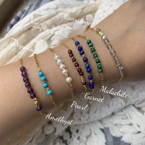 Könnte beinhalten: Ein Set aus neun goldenen Kettenarmbändern mit unterschiedlich farbigen Edelsteinperlen. Die Armbänder sind mit den Namen der Edelsteine beschriftet: Amethyst, Türkis, Lapislazuli, Perle, Granat, Malachit, Mondstein.