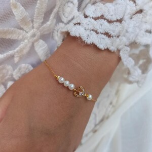 Peut inclure: Bracelet en or orné d'un charm en forme de cœur serti de petits cristaux transparents et d'une rangée de perles blanches. Le bracelet est porté sur un poignet, avec une manche en dentelle blanche en arrière-plan.