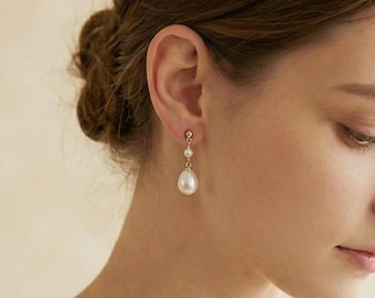 May include: Gold-toned drop earrings featuring a small round pearl and a larger, teardrop-shaped pearl. The earrings are shown on a person's ear, highlighting the elegant design and classic style. The pearls have a lustrous, white appearance.