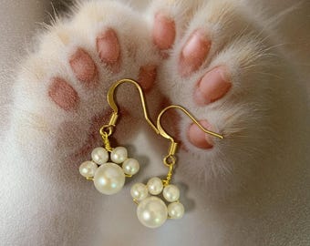 May include: A pair of gold-colored earrings shaped like cat paws, each composed of several white, round pearls. The earrings are suspended from gold-colored hooks. The background features a blurred image of a cat's paw.