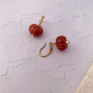 May include: A pair of gold-colored earrings featuring small, carved, reddish-orange pumpkin charms. The pumpkins have detailed ridges and are attached to gold-colored ear wires. The earrings are displayed on a textured, off-white surface.