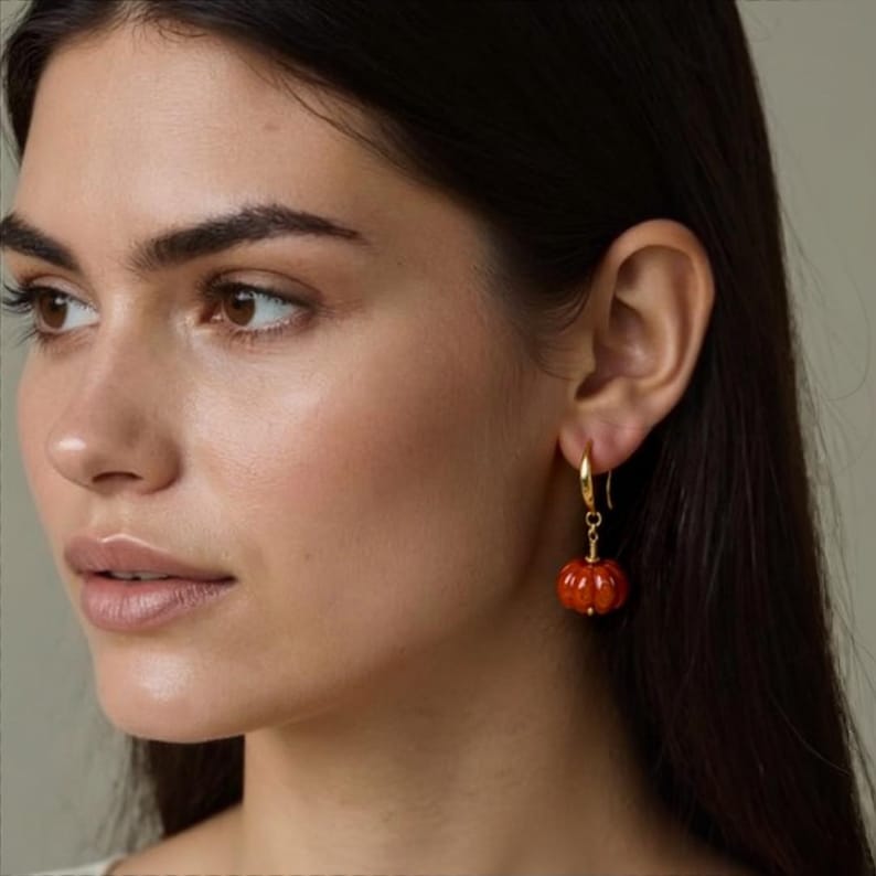 May include: Gold hoop earrings with a dangling, carved orange pumpkin charm. The pumpkin has a textured surface and is suspended from a delicate gold chain. The earrings are a fall-themed accessory.