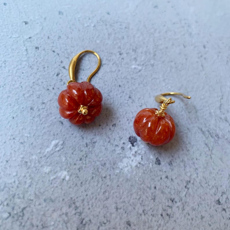 May include: A pair of gold-colored earrings, each featuring a carved, reddish-orange pumpkin design. The pumpkins have a detailed, textured surface and are suspended from gold-colored hooks, creating a festive autumn theme.