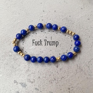 May include: A beaded bracelet featuring deep blue lapis lazuli beads and gold-colored spacer beads. The text "Fuck Trump" is written in black letters within a dotted oval shape.