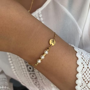 Könnte beinhalten: Ein goldenes Kettenarmband mit einem kleinen runden Anhänger mit dem Buchstaben "S" und einer Reihe weißer Perlen.