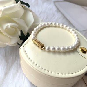 Könnte beinhalten: Ein weißes Perlenarmband mit einem goldenen Anhänger, auf dem "Sebastian Charm" steht. Das Armband befindet sich in einer weißen Box mit einem goldenen Verschluss.