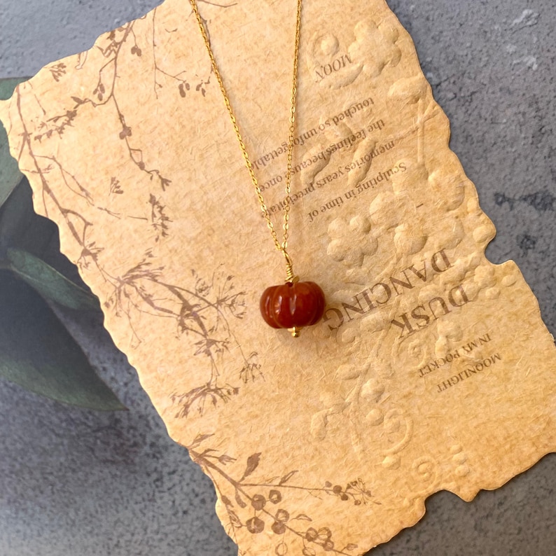 May include: A gold-colored necklace with a small, reddish-brown pumpkin pendant. The pendant is intricately detailed and hangs from a delicate chain. The necklace rests on a textured, tan paper with the words "Dusk Dancing" and "Moonlight".