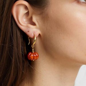 Peut inclure: Boucle d'oreille dorée avec un charm en forme de citrouille. Le charm est de couleur orange vif avec des rainures verticales, et la boucle d'oreille est fixée à l'oreille par un crochet doré. Accessoire sur le thème de l'automne.