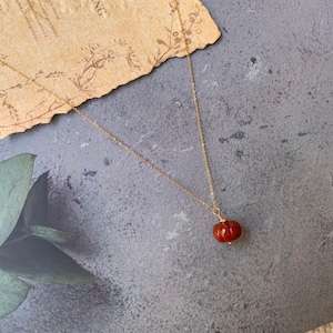 May include: A gold-colored necklace with a small, red, pumpkin-shaped pendant. The pendant has a textured surface and a gold-colored cap. The necklace is displayed on a gray surface with a piece of aged paper in the background.