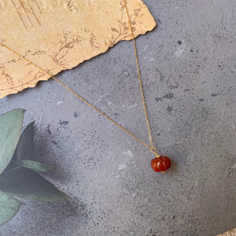 May include: A gold-colored necklace with a small, red, pumpkin-shaped pendant. The pendant has a textured surface and a gold-colored cap. The necklace is displayed on a gray surface with a piece of aged paper in the background.