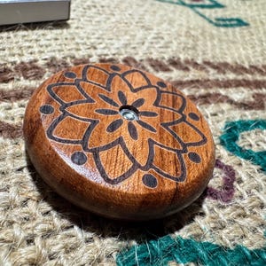 Peut inclure: Un objet circulaire en bois avec une finition polie et un motif mandala gravé au laser en brun foncé sur sa surface supérieure. Le centre du motif présente un petit insert métallique circulaire, créant un contraste entre le bois et le métal.